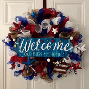 K&ouml;nnte beinhalten: Ein patriotischer Kranz mit rotem, wei&szlig;em und blauem Mesh, Sternen und Akzenten. Ein blaues Holzschild in der Mitte tr&auml;gt die Aufschrift "Welcome" und "God Bless My Home". Der Kranz ist f&uuml;r eine Haust&uuml;r gedacht.