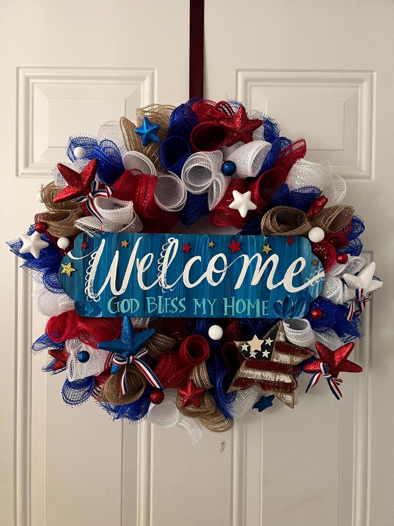 K&ouml;nnte beinhalten: Ein patriotischer Kranz mit rotem, wei&szlig;em und blauem Mesh, Sternen und Akzenten. Ein blaues Holzschild in der Mitte tr&auml;gt die Aufschrift "Welcome" und "God Bless My Home". Der Kranz ist f&uuml;r eine Haust&uuml;r gedacht.