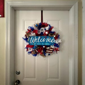 K&ouml;nnte beinhalten: Ein patriotischer Kranz mit rotem, wei&szlig;em und blauem Netz, Sternen und B&auml;ndern, der an einer wei&szlig;en T&uuml;r h&auml;ngt. Ein blaues Schild in der Mitte lautet "Welcome God Bless My Home."
