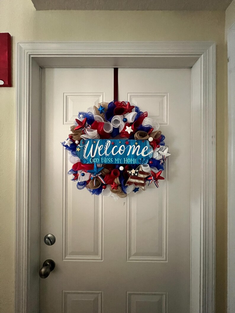 K&ouml;nnte beinhalten: Ein patriotischer Kranz mit rotem, wei&szlig;em und blauem Netz, Sternen und B&auml;ndern, der an einer wei&szlig;en T&uuml;r h&auml;ngt. Ein blaues Schild in der Mitte lautet "Welcome God Bless My Home."