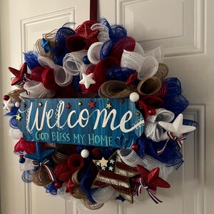 K&ouml;nnte beinhalten: Ein patriotischer Kranz aus rotem, wei&szlig;em und blauem Netz, Sternen und dekorativen Elementen. Ein blaues Schild in der Mitte tr&auml;gt die Aufschrift "Welcome, God Bless My Home".