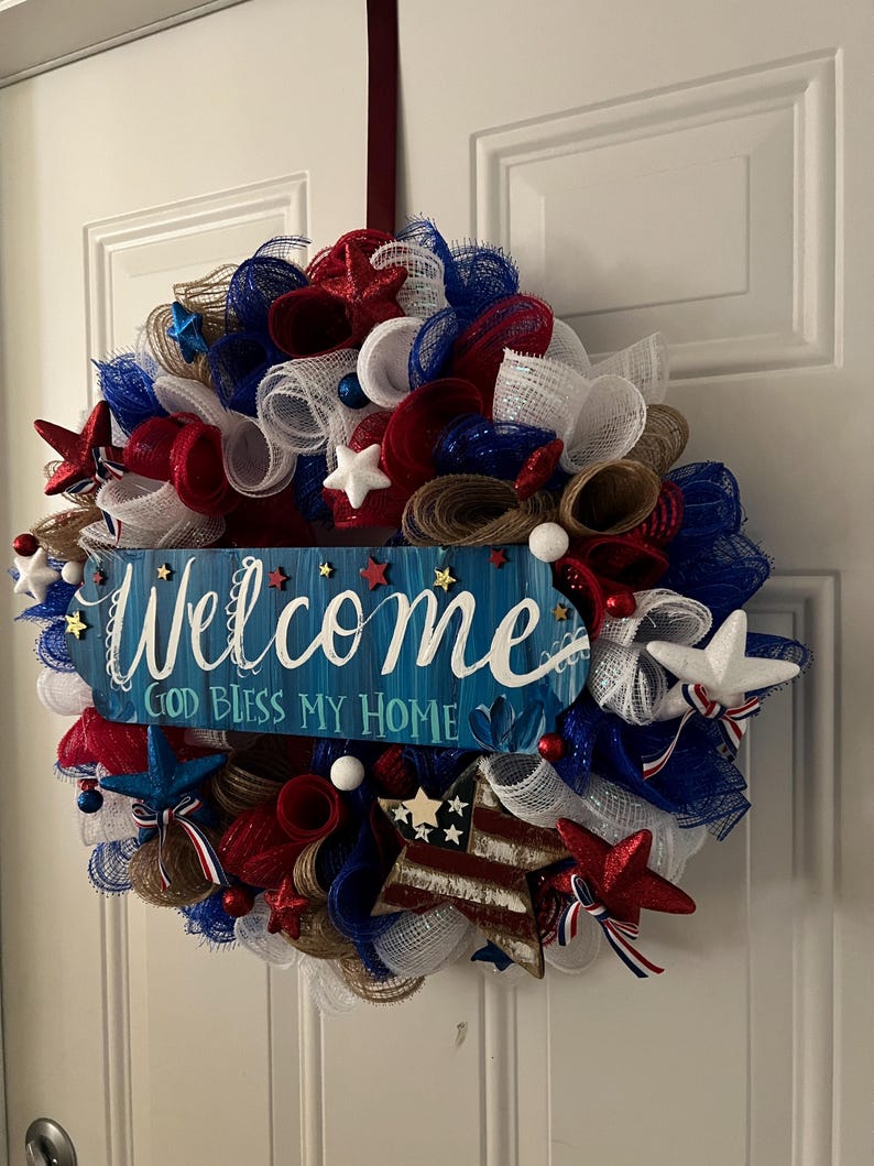 K&ouml;nnte beinhalten: Ein patriotischer Kranz aus rotem, wei&szlig;em und blauem Netz, Sternen und dekorativen Elementen. Ein blaues Schild in der Mitte tr&auml;gt die Aufschrift "Welcome, God Bless My Home".