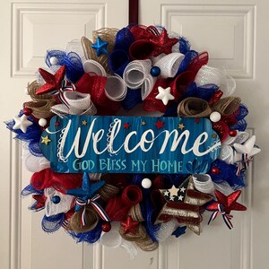 K&ouml;nnte beinhalten: Ein patriotischer Kranz mit rotem, wei&szlig;em und blauem Netz, Sternen und Akzenten. Ein blaues Holzschild in der Mitte tr&auml;gt die Aufschrift "Welcome, God Bless My Home" in wei&szlig;er Schrift. Der Kranz h&auml;ngt an einer wei&szlig;en T&uuml;r.
