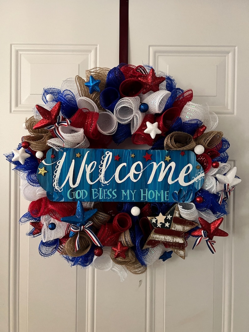 K&ouml;nnte beinhalten: Ein patriotischer Kranz mit rotem, wei&szlig;em und blauem Netz, Sternen und Akzenten. Ein blaues Holzschild in der Mitte tr&auml;gt die Aufschrift "Welcome, God Bless My Home" in wei&szlig;er Schrift. Der Kranz h&auml;ngt an einer wei&szlig;en T&uuml;r.