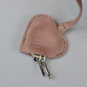 Puede incluir: Un llavero de cuero rosa en forma de corazón con la palabra "MOSCHINO" grabada en relieve. El llavero tiene un anillo de llaves plateado con dos llaves.