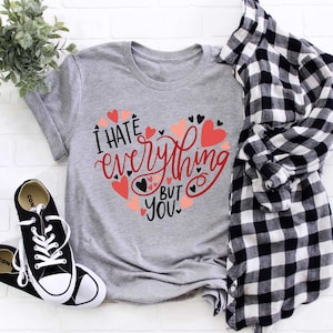 Puede incluir: Camiseta gris con un diseño en forma de corazón que dice "Odio todo menos a ti". El corazón está lleno de corazones más pequeños en negro y rosa.
