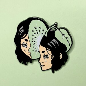 May include: Black and green enamel pin featuring two stylized faces with a kiwi fruit design. The kiwi fruit is split in half, with one face visible through the fruit.