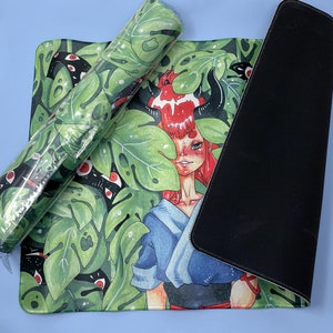 May include: A large, colorful mousepad with a black border. The design features a woman with red hair and horns, surrounded by green leaves and black snakes with red eyes. The woman is wearing a blue and black outfit.