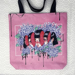 May include: Pink tote bag with black straps featuring a graphic design of a woman's face with multiple eyes surrounded by purple flowers. The design has a dripping black paint effect.