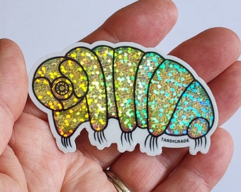 May include: A holographic sticker of a tardigrade, also known as a water bear, held in a hand. The sticker has a segmented body with iridescent glitter, and the word "TARDIGRADE" is printed on the bottom.