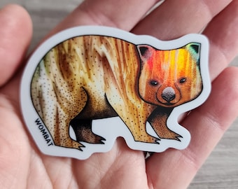 Puede incluir: Una colorida pegatina con una ilustración de un wombat. El wombat tiene un cuerpo marrón con un patrón texturizado y una cabeza de colores del arcoíris. La palabra "WOMBAT" está impresa en negro en la pegatina.