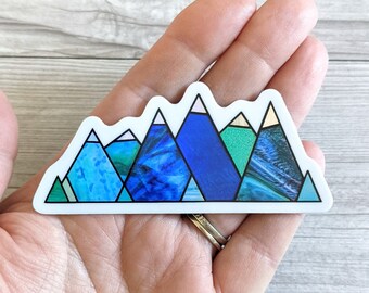 May include: A mountain range sticker with a white border. The sticker features a series of stylized mountain peaks in shades of blue, teal, and green, with white and gold accents. The design is geometric and abstract.