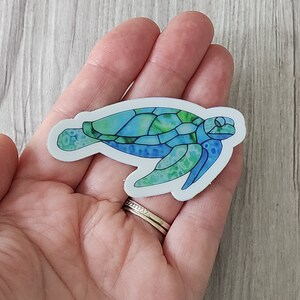 May include: A sticker featuring a sea turtle design. The turtle has a blue and green stained glass effect, with a white border. The sticker is held in a hand, with a light wood grain background.