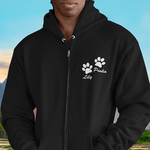 May include: Black zip-up hoodie with white paw prints and the names "Lily" and "Pooba" printed above the paw prints. The hoodie has a drawstring hood and two front pockets. The background shows a lake and mountains.