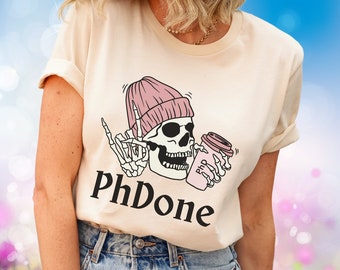 May include: Cream-colored t-shirt featuring a graphic of a skeleton wearing a pink beanie and holding a coffee cup. The skeleton is giving the middle finger. The text "PhDone" is printed below the graphic.