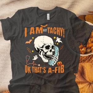 May include: A dark gray t-shirt with a white skull holding a blue phone and the text "I AM NOT TACHY! OK THAT'S A-FIB" with a stethoscope, a heart rate, and other Halloween-themed graphics.
