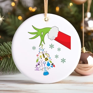May include: A white ceramic Christmas ornament featuring a green hand with a red Santa hat holding a Christmas tree made of science-themed icons. The tree is surrounded by snowflakes.