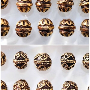 Könnte beinhalten: Eine Sammlung von 20 goldfarbenen Metallperlen mit komplizierten Designs. Die Perlen sind in zwei Reihen zu je zehn angeordnet, wobei jede Perle ein einzigartiges Muster aufweist.