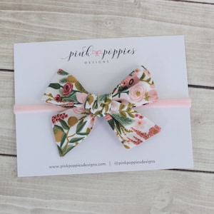 K&ouml;nnte beinhalten: Ein rosa Haarband mit einem floralen Muster in Rosa. Die Schleife ist in einem Knoten gebunden und hat einen wei&szlig;en Hintergrund mit rosa, roten und gr&uuml;nen Blumen. Der Text "pink poppies designs" ist auf einer wei&szlig;en Karte hinter der Schleife gedruckt.