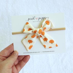 May include: A cream-colored baby bow with a repeating pattern of orange pumpkins. The bow is attached to a tan headband and displayed on a white card with the text "pink poppies designs".