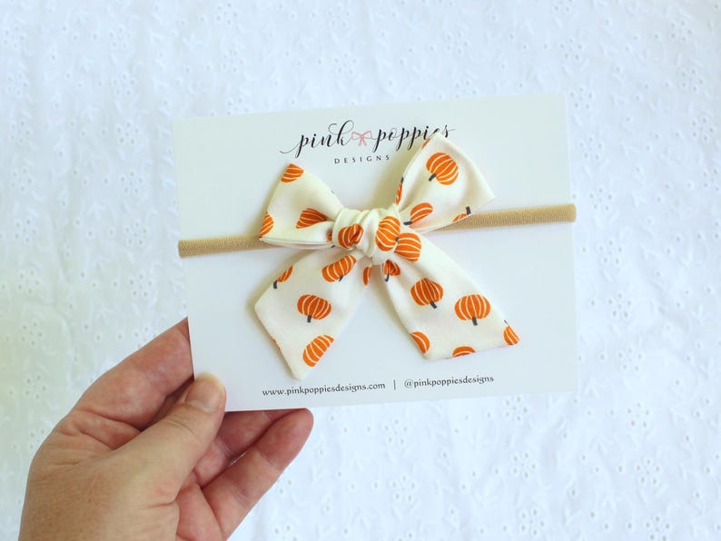 May include: A cream-colored baby bow with a repeating pattern of orange pumpkins. The bow is attached to a tan headband and displayed on a white card with the text "pink poppies designs".