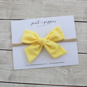 May include: A yellow fabric bow headband with a nude elastic band. The headband is on a white card with the text "pink poppies designs" and a website address.