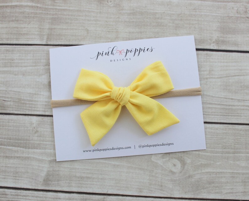 May include: A yellow fabric bow headband with a nude elastic band. The headband is on a white card with the text "pink poppies designs" and a website address.