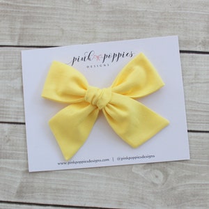 May include: A bright yellow fabric bow with a center knot. The bow is on a white card with the text "pink poppies designs" and website information.