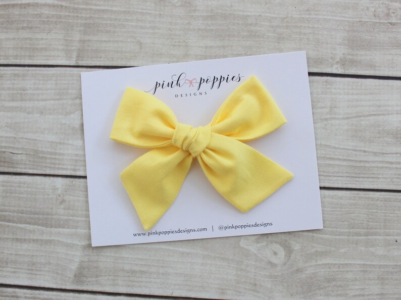 May include: A bright yellow fabric bow with a center knot. The bow is on a white card with the text "pink poppies designs" and website information.