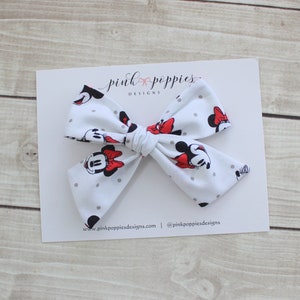 May include: White bow with a Minnie Mouse print and gray polka dots. The bow is tied with a red ribbon. The bow is on a white card with the text "pink poppies designs" and a website address.