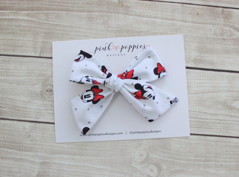 May include: White bow with a Minnie Mouse print and gray polka dots. The bow is tied with a red ribbon. The bow is on a white card with the text "pink poppies designs" and a website address.