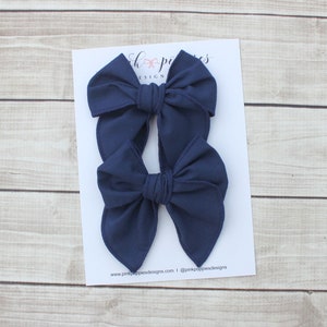 May include: Two navy blue fabric hair bows. The bows are tied in a simple knot and have a slightly pointed shape. The bows are on a white background with the text "Pink Poppies Designs" and a website address.