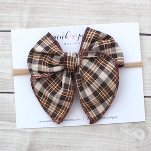 Peut inclure: Un grand nœud papillon à carreaux marron, composé de marron, beige et crème, fixé sur un bandeau beige. Le nœud est posé sur une carte blanche avec le texte "pink po designs" et une adresse de site web. Le motif du nœud est à carreaux classiques.