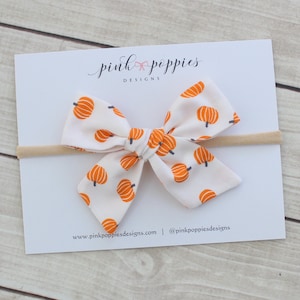Può includere: Un fiocco di tessuto bianco con un motivo ripetuto di zucche arancioni, attaccato a una fascia beige. Il fiocco è centrato su una carta bianca con il testo "pink poppies designs" e informazioni sul sito web.