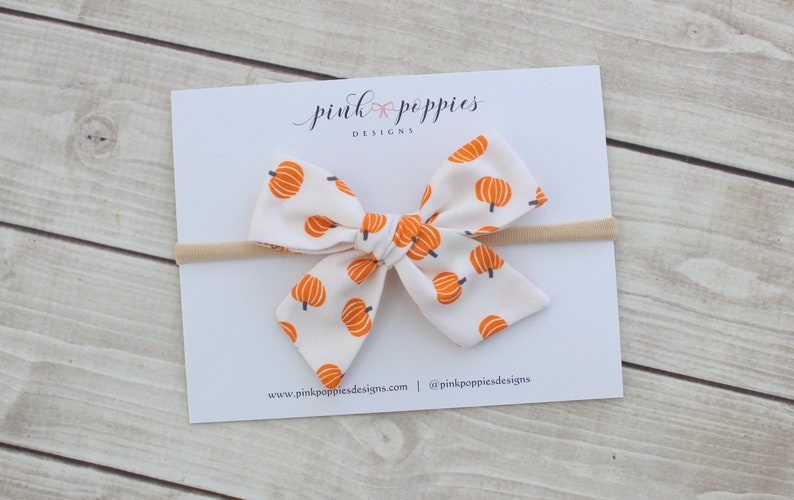 May include: A white fabric bow with a repeating orange pumpkin pattern, attached to a beige headband. The bow is centered on a white card with the text "pink poppies designs" and website information.