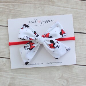 May include: A white headband with a red elastic band and a white bow with a Minnie Mouse print. The bow has gray polka dots and features Minnie Mouse's face with red bows. The headband is labeled "pink poppies designs".