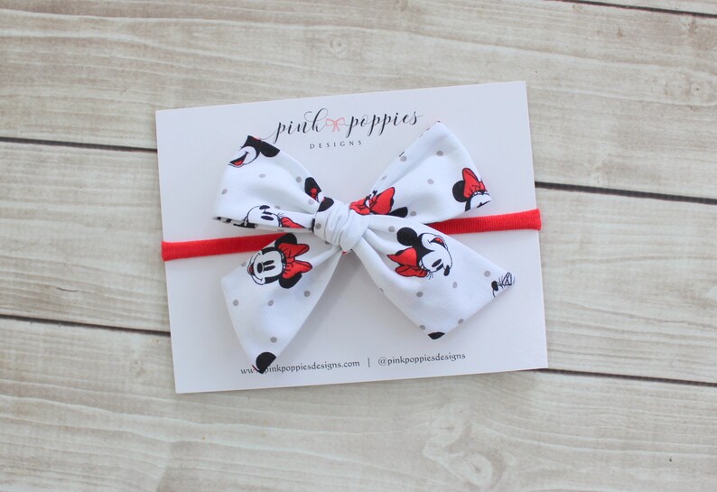 May include: A white headband with a red elastic band and a white bow with a Minnie Mouse print. The bow has gray polka dots and features Minnie Mouse's face with red bows. The headband is labeled "pink poppies designs".