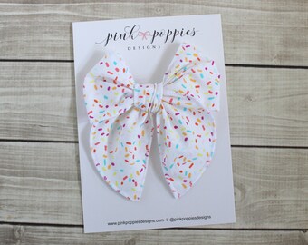Puede incluir: Un lazo blanco con un estampado de confeti en tonos de rosa, naranja, amarillo, azul y verde. El lazo está sobre una tarjeta blanca con el texto "pink poppies designs" y una dirección de sitio web.