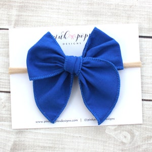 May include: A royal blue fabric bow headband with a nude elastic band. The bow is tied in a classic style with a pointed tip on each side. The headband is on a white card with the text "pink poppies designs" and a website address.