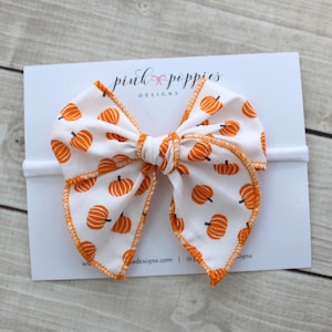 Könnte beinhalten: Weißes Stoff-Haarband mit einem orangefarbenen Kürbismuster und orangefarbenen Nähten. Die Schleife ist an einem weißen Stirnband befestigt. Der Text "pink poppies DESIGNS" ist auf die weiße Karte gedruckt.