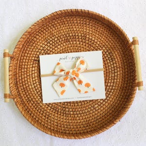 May include: A white and orange pumpkin print bow on a headband, displayed on a white card. The card is on a round, woven tray with bamboo handles. The bow features a repeating pattern of orange pumpkins.
