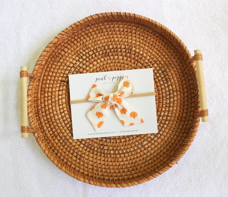 May include: A white and orange pumpkin print bow on a headband, displayed on a white card. The card is on a round, woven tray with bamboo handles. The bow features a repeating pattern of orange pumpkins.