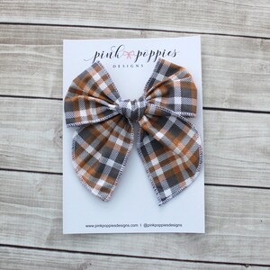 May include: A large hair bow with a plaid pattern in shades of brown, gray, and white, displayed on a white card. The card features the text "pink poppies DESIGNS" and a website address. The bow is centered on a light wood grain surface.