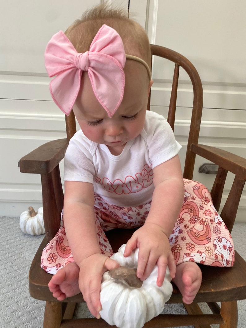 May include: A baby girl wearing a white shirt with pink lettering, pink and white patterned pants, and a pink bow headband. The baby is sitting in a wooden rocking chair and holding a white pumpkin.