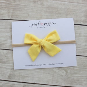 May include: A yellow fabric bow headband with a nude elastic band. The headband is on a white card with the text "pink poppies designs" and website information.