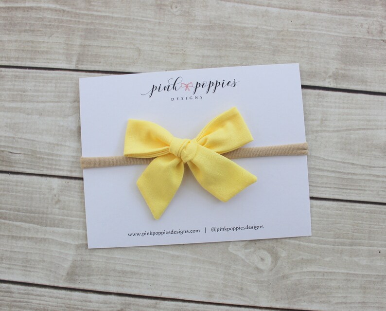 May include: A yellow fabric bow headband with a nude elastic band. The headband is on a white card with the text "pink poppies designs" and website information.