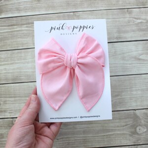 May include: A large pink fabric bow with a double loop design. The bow is on a white card with the text "pink poppies designs" printed on it.