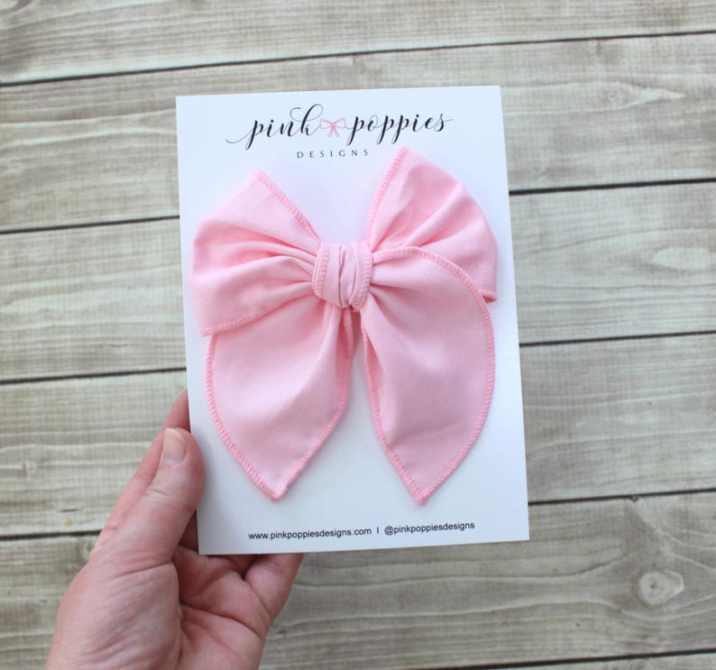 May include: A large pink fabric bow with a double loop design. The bow is on a white card with the text "pink poppies designs" printed on it.