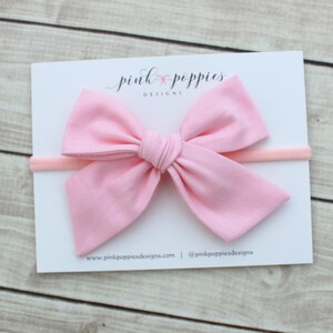 May include: A pink fabric bow headband with a white card behind it. The card has the text "pink poppies designs" on it.