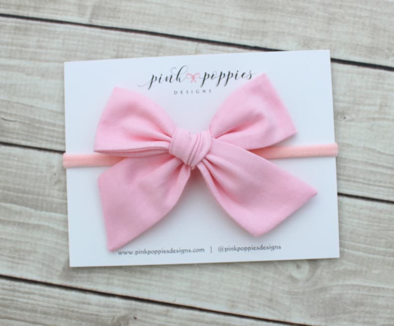 May include: A pink fabric bow headband with a white card behind it. The card has the text "pink poppies designs" on it.